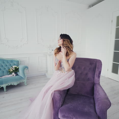 Beautiful woman model in a wedding dress sit on a chair in the spacious room. Young attractive lady in a beautiful evening dress of lavender color with pebbles color sits armchair.