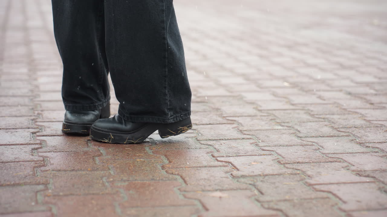 Foot of person wearing black trousers and black boots turning on wet ground, creating ripples on surface, highlighting motion in urban setting after rain, walking on pavement or outdoor surface