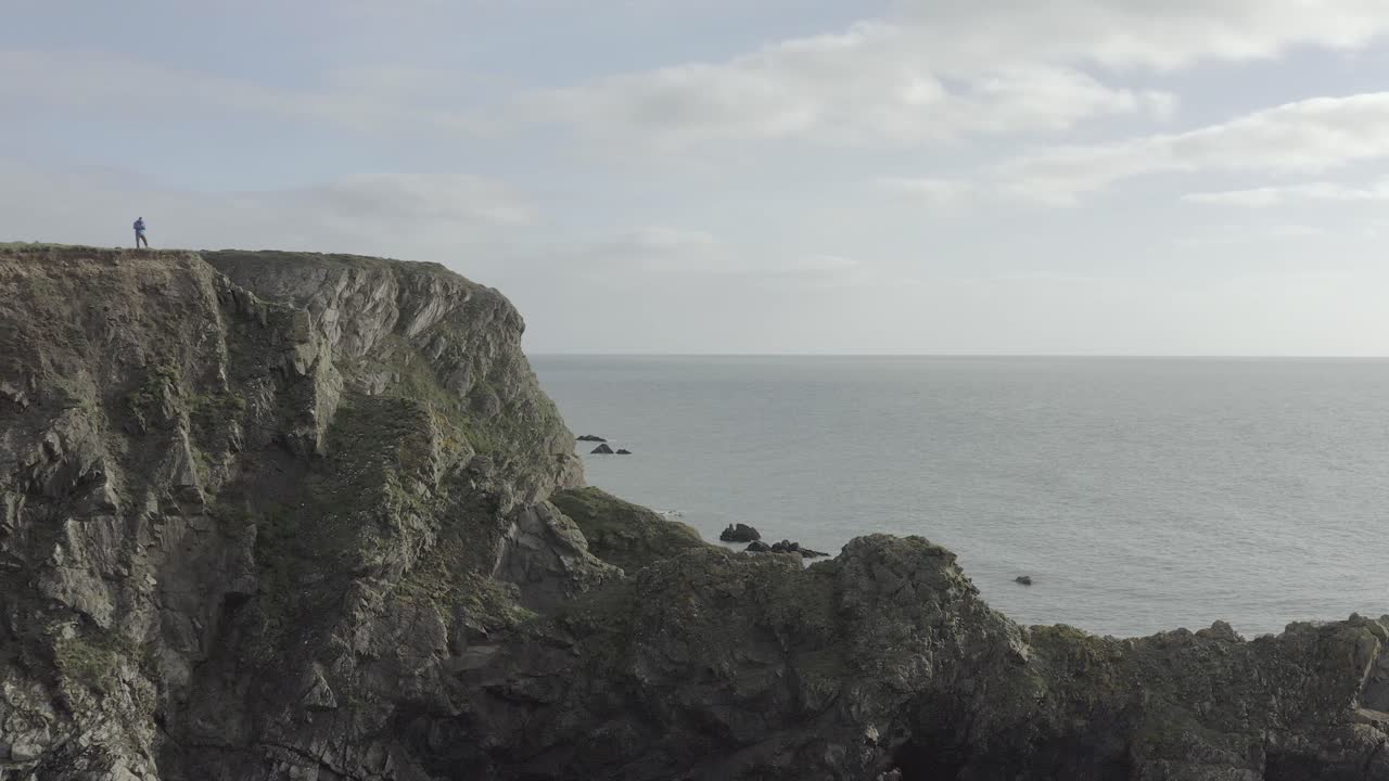 Static shot of drone pilot atop steep rock cliffs overlooking ocean