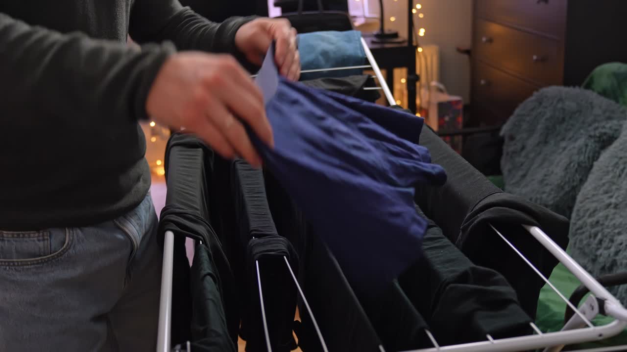 Man hanging navy briefs on an indoor drying rack, surrounded by dark clothes in a cozy apartment lit by warm fairy lights