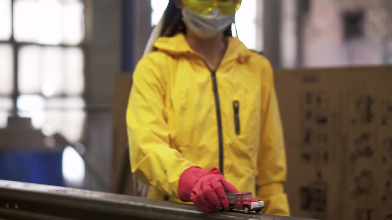 una niña positiva con ropa de trabajo protectora - chaqueta amarilla, gafas, guantes y máscara en el centro de reciclaje. imágenes de cerca de una niña jugando con un coche de juguete, empujarlo por la barandilla
