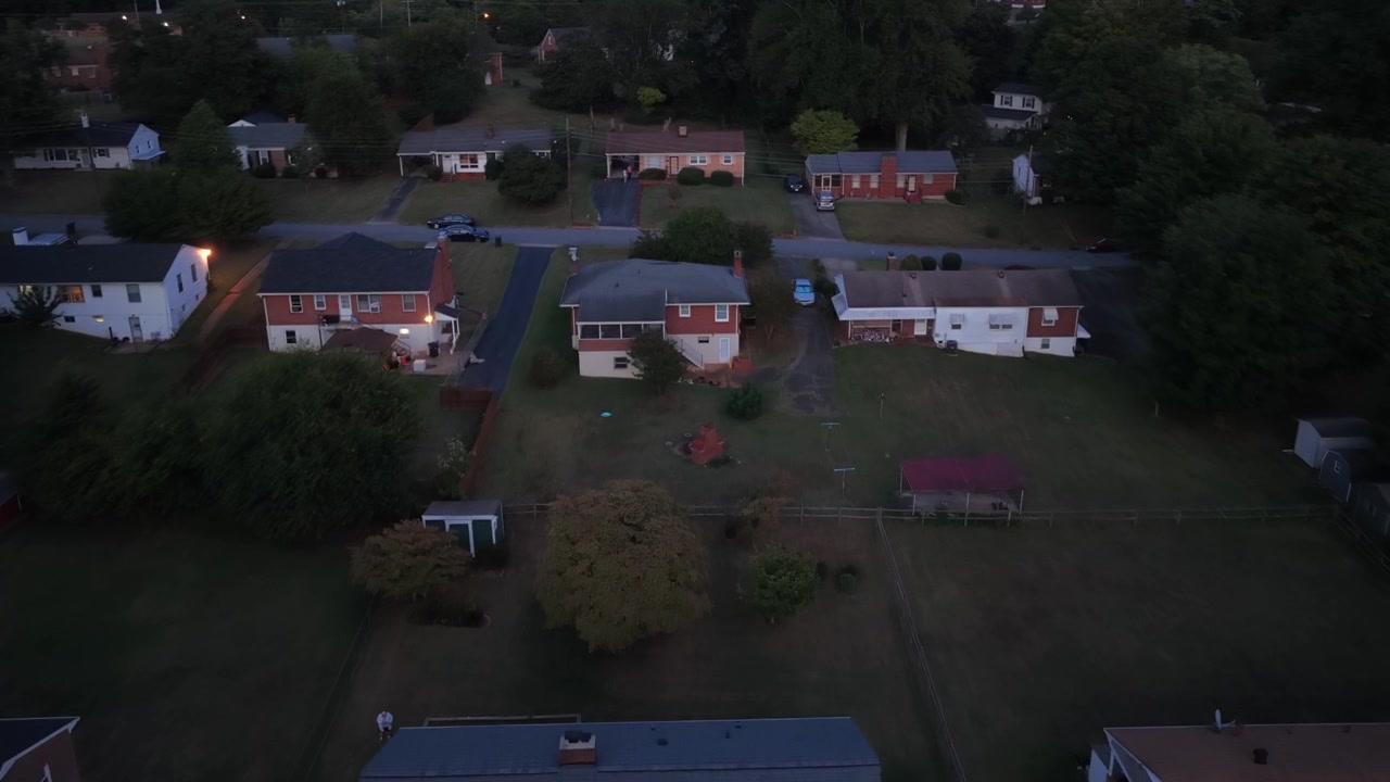 Brick ranch houses stand in rows along quiet cross streets in Lynchburg, Virginia, at dusk, with carports, parked cars and wide grass yards between them. Aerial flyover shot