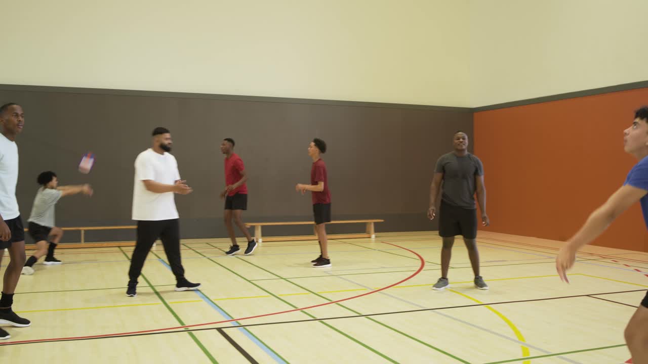 Diverse male teammates practicing rally in gym after back player tossing purple ball for passing