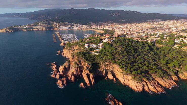 Stunning aerial perspective flying over the rugged cliffs toward the coastal town of sant feliu de guixols in costa brava, spain, capturing the marina and beach at sunrise