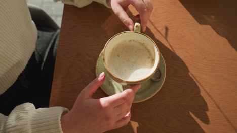 overhead view of person in white sweater playfully turning ceramic tea cup on wooden cafe table, warm sunlight casting shadows across scene, relaxed mood with visible spoon