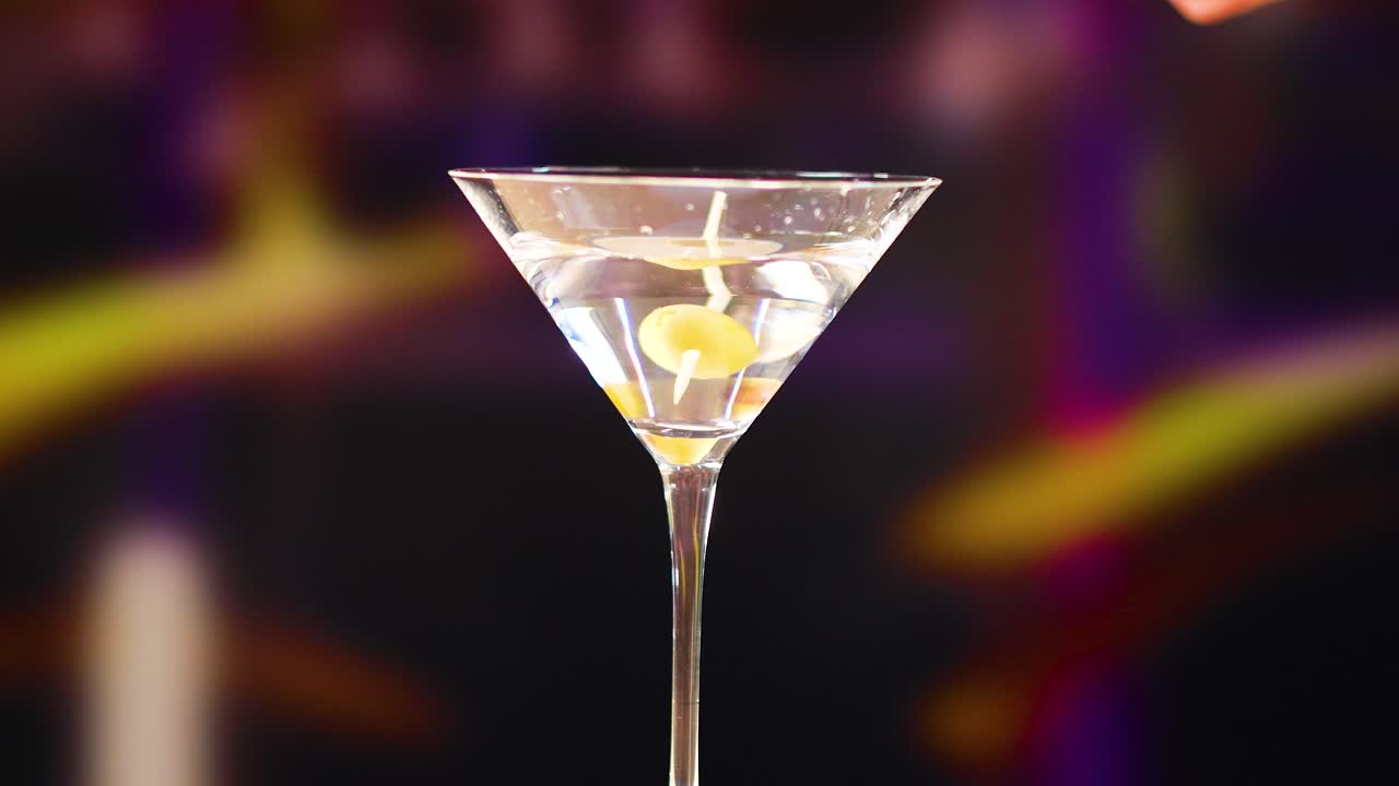 A martini glass is garnished with olives in a dimly lit bar. The background is blurred, highlighting the drink