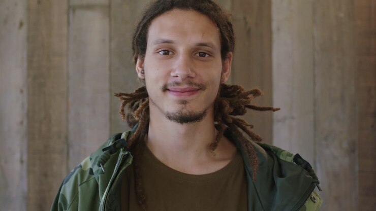 ritratto ravvicinato di un giovane attraente uomo di razza mista coi capelli dreadlocks sorridente guardando la telecamera uomo sicuro di sé che indossa una giacca di camuffamento sfondo di legno