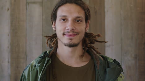 close up portrait of young attractive mixed race man dreadlocks hairstyle smiling looking at camera confident male wearing camouflage jacket wooden background