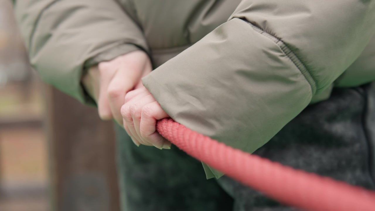 Closeup hands gripping red rope framed by puffy green jacket, knuckles tensed, slow pull across playground post, autumn light, tactile rope strands, intimate study of effort and focus, cinematic