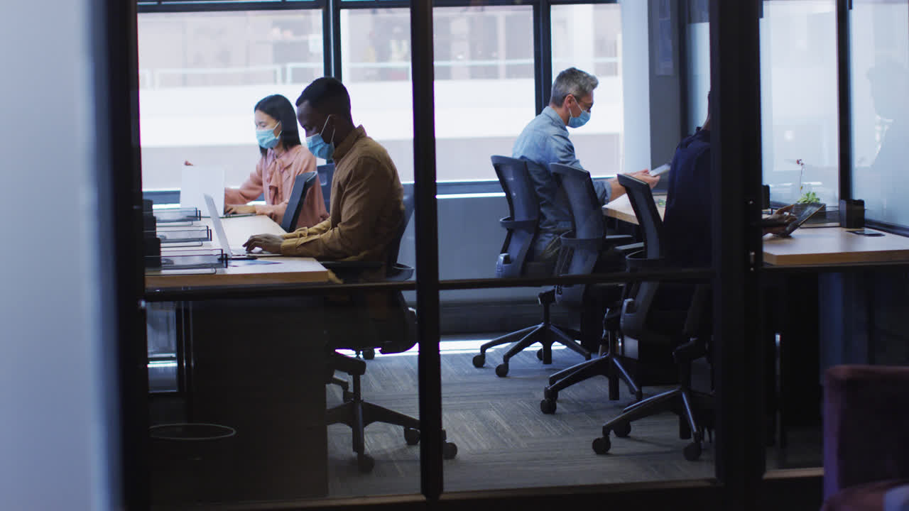 현대적인 사무실에서 책상에 앉아 일하는 사무실 동료들이 얼굴 마스크를 착용하고 있습니다.