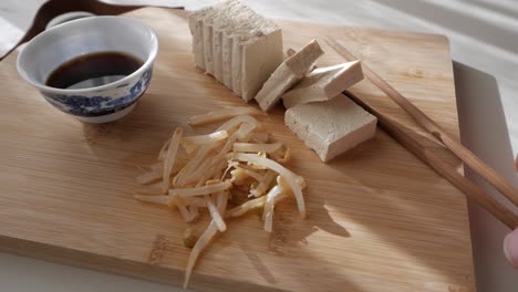 Sliced tofu with bean sprouts, soy sauce, and chopsticks on a wooden board