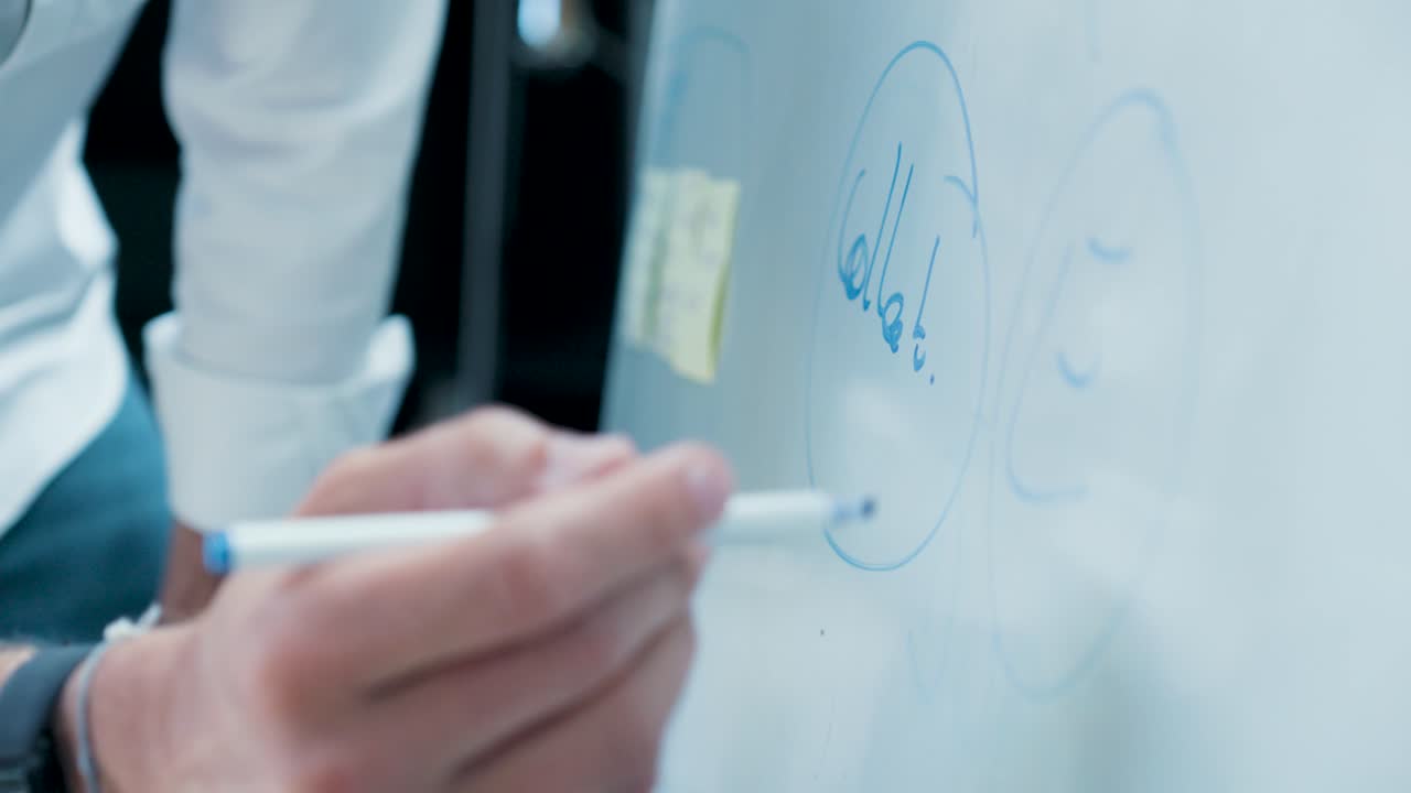 Close-up on a whiteboard, a man in a white shirt with a wristband and watch writes on the board with a blue felt-tip pen.