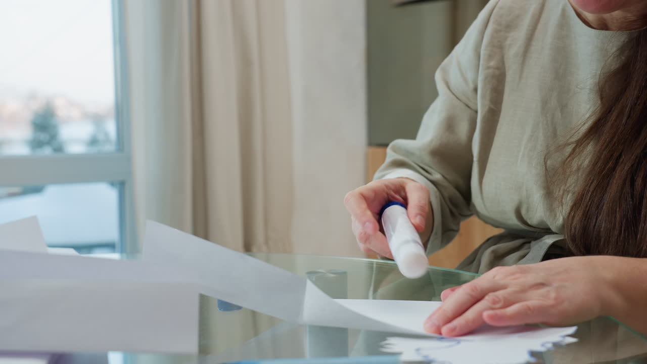 Hand view of woman in gray gown applying glue on paper craft, focus on hands using glue stick for crafting in cozy home setting with snow outside window in background