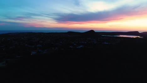 Wide-spread-blue-hour-dusk-glow-sunset-across-ocean-above-calm-port-town,-aerial