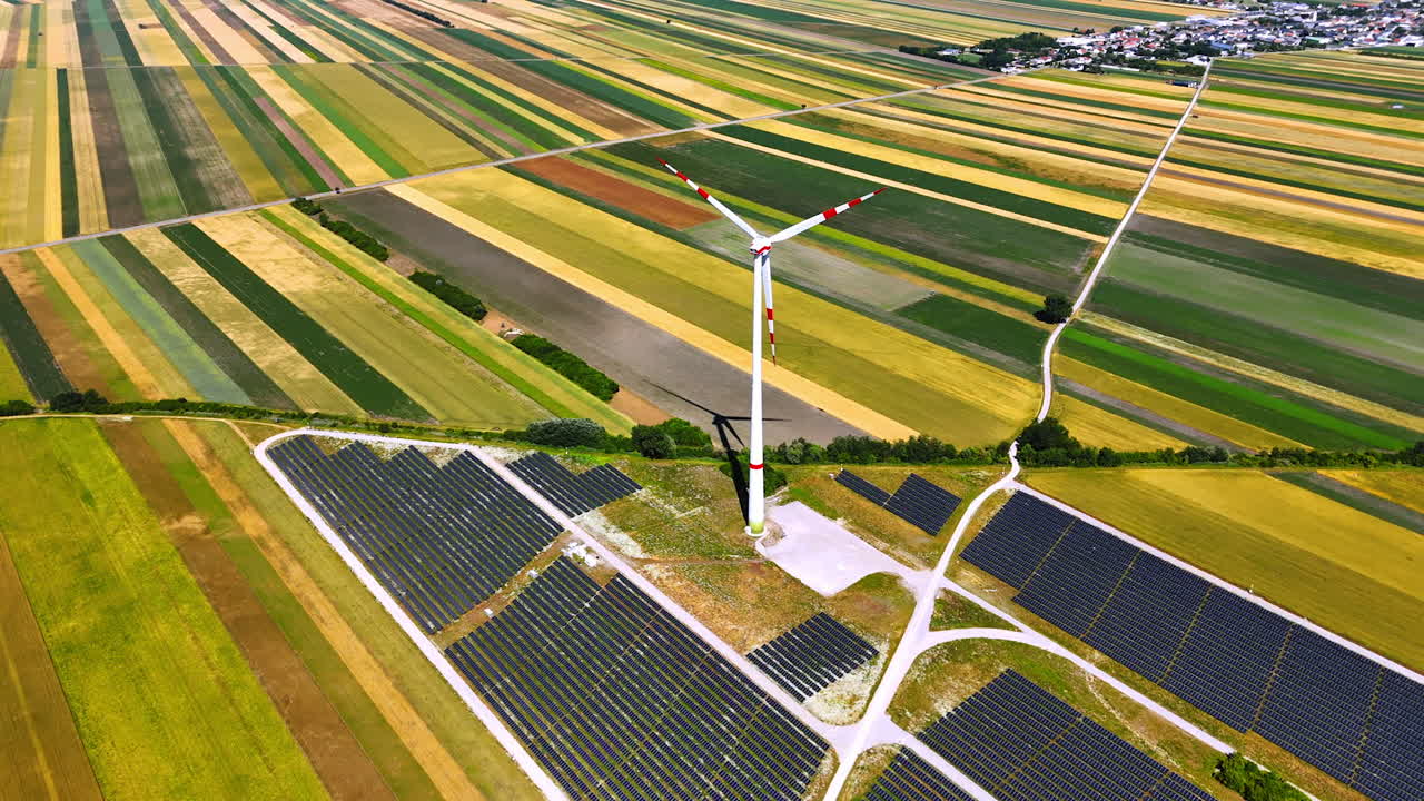 Multiple solar panels surround the wind turbine. Drone footage over the colorful fields on sunny day. Village at backdrop