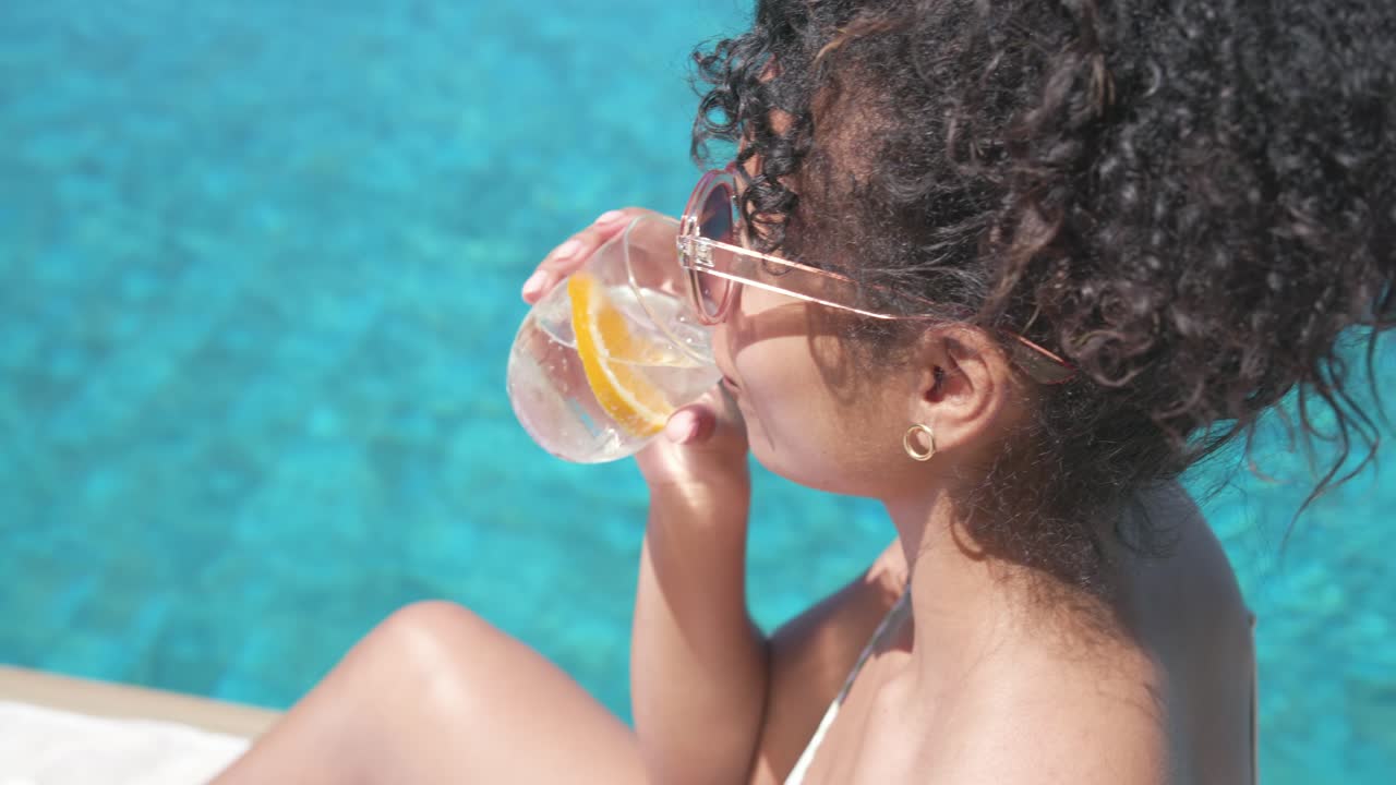 Woman is lowering clear sunglasses as sunlight is warming face, sipping water with orange slice