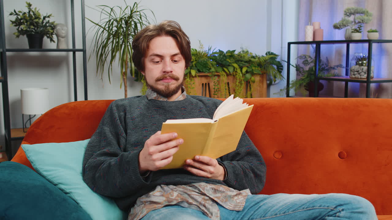 Bärtiger Mann liest ein interessantes Buch, blättert die Seiten, lächelt, genießt Literatur, ruht sich zu Hause aus