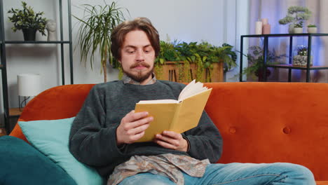 bearded man reading interesting book turning pages smiling enjoying literature taking a rest at home