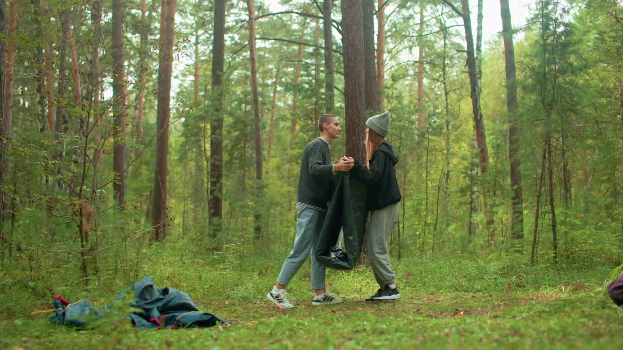 Man and wife hold opposite sides of tent fabric while packing up in forest, briefly leaning in for kiss, surrounded by trees and scattered gear, sharing affectionate moment during camping departure
