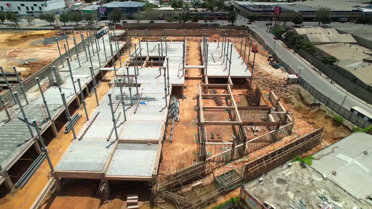 Tomada aérea simétrica de drone voando para trás e ascendendo sobre um canteiro de obras de um edifício comercial, revelando lajes de concreto, colunas de aço e a cidade circundante.