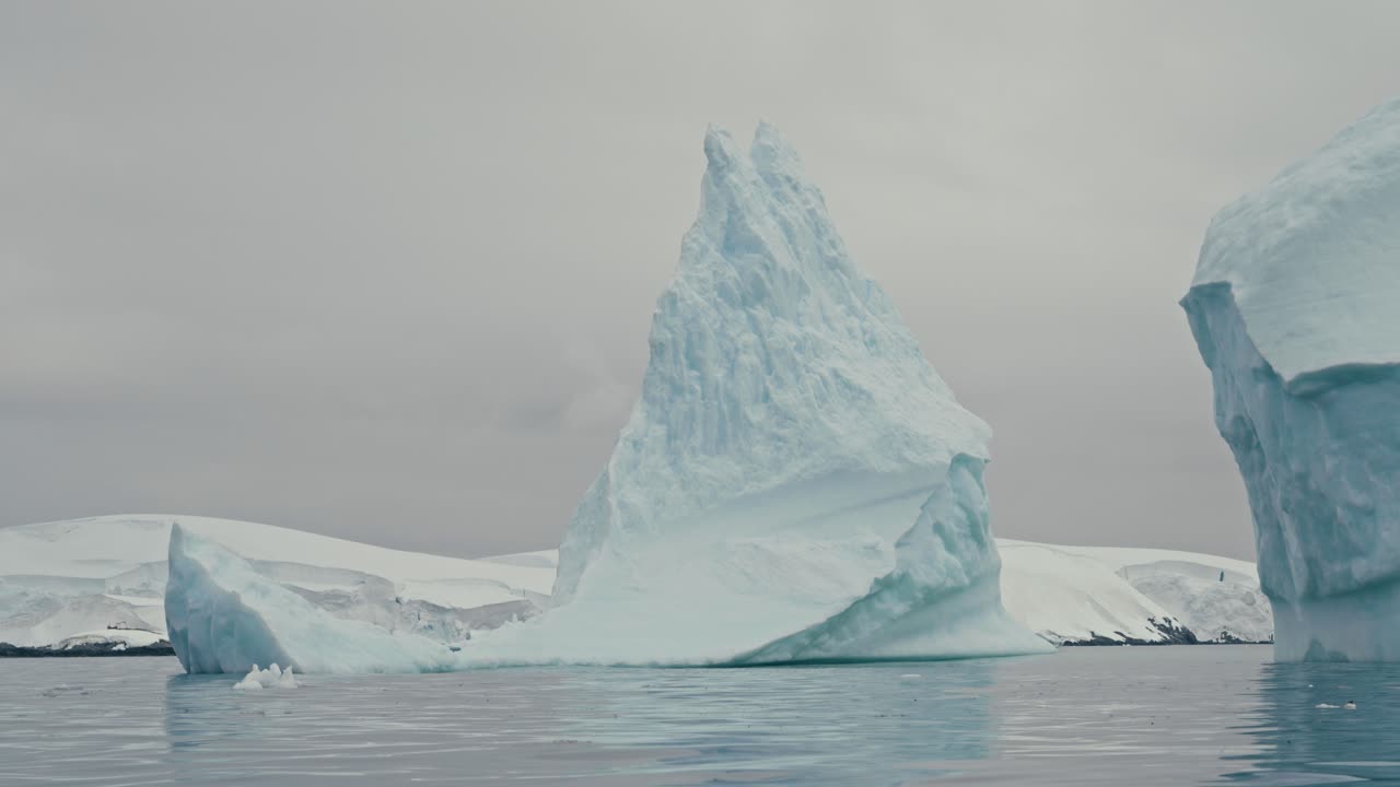 Eterisk långsam panorering förbi ett högt, spetsigt isberg och en bredare eroderad formation som flyter på glasklara, lugna antarktiska polarvatten under en platt grå mulen himmel.
