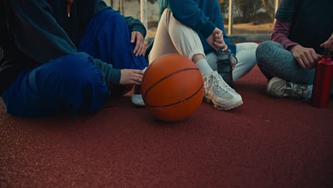 Close-up-three-girls-in-sportswear-are-holding-sports-bottles-and-sitting-on-the-red-floor-near-an-orange-ball-on-a-basketball-street-court-in-the-morning