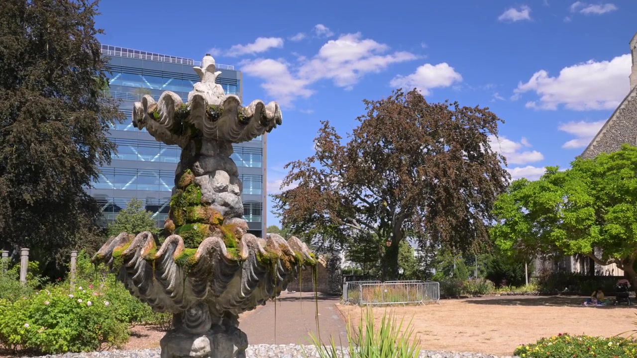 Un'antica fontana in pietra si erge nei Forbury Gardens, Reading, incorniciata da alberi e moderni uffici in vetro sotto un cielo azzurro brillante