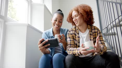 Happy-diverse-couple-sitting-on-stairs,-drinking-coffee-using-smartphone-and-talking,-slow-motion