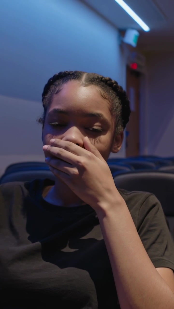 Vertical video: Yawning teen in black tee lifting right hand to mouth in padded seats stifling yawn