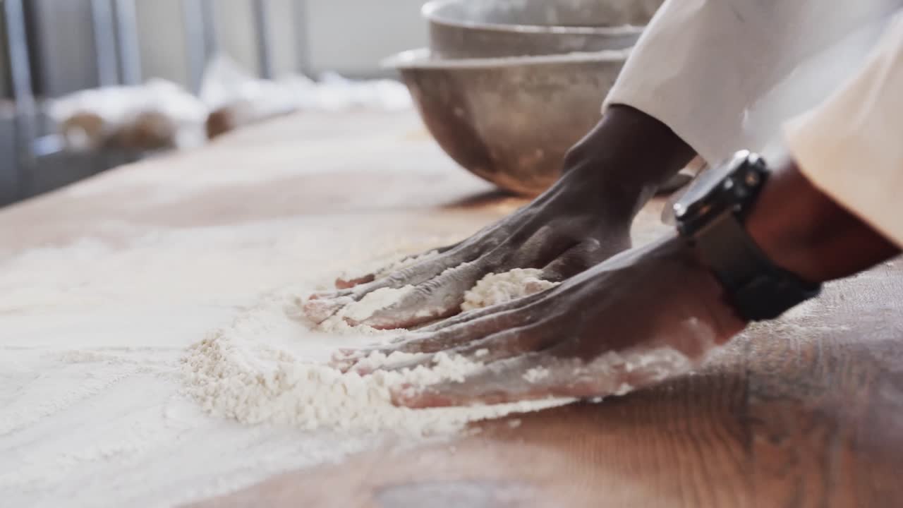 African american male baker working in bakery kitchen, spreading flour on counter in slow motion