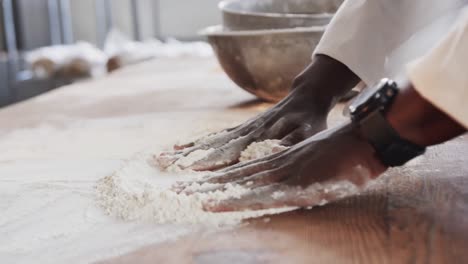 african american male baker working in bakery kitchen, spreading flour on counter in slow motion