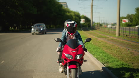 two ladies ride a power bike wearing helmets close to a railway track, a blur view of a car with its headlamp on coming behind, while electric poles and signposts are visible in the background