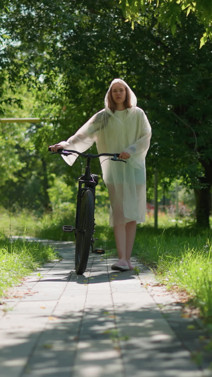 반투명한 레인코트를 입은 여성이 자전거를 타고 은 길을 고, 울창한 초록색과 나무로 둘러싸여 있으며, 건물과 다른 사람이 배경에서 고 있습니다.