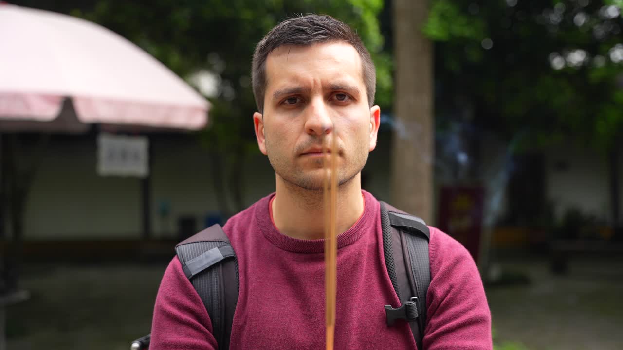 Primer plano de un hombre turista mirando seriamente a la cámara mientras quema varitas de incienso.