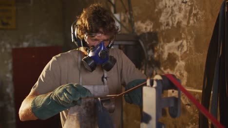 caucasian male blacksmith wearing breathing mask and ear guards, forging metal tool in workshop