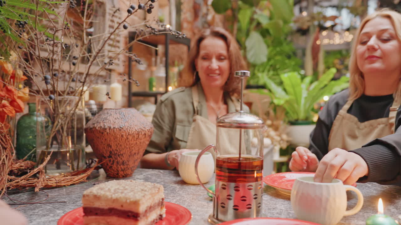 White florist served homemade cake slice, barista in green apron brings red plate to florists at plant shop counter, french press tea, ceramic cup, rustic decor, warm chat, customer interaction