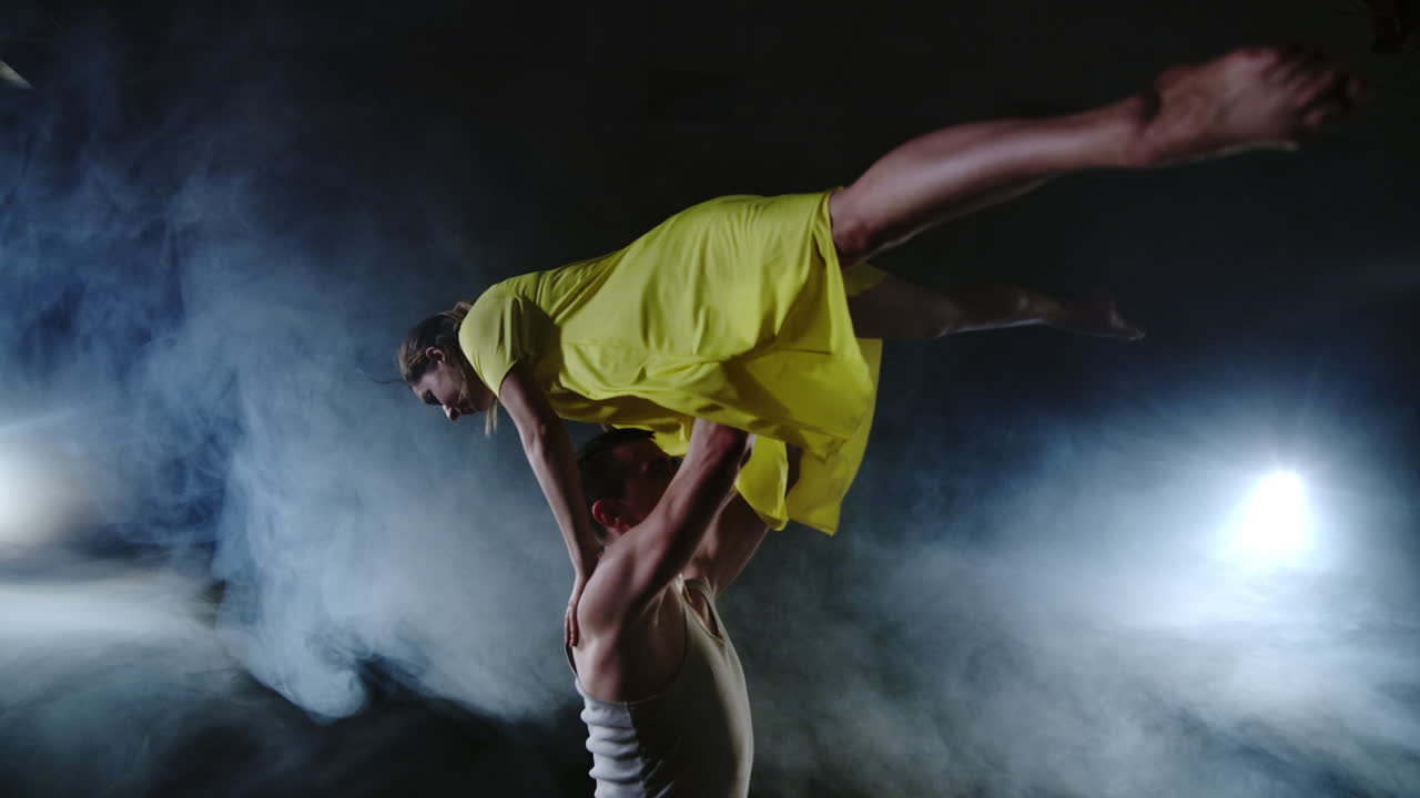 Two dancers a man and a woman run to each other and a male partner raises a woman in a yellow dress in her arms and rotates in the air performing top support.