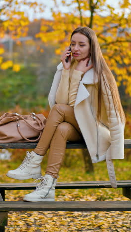 attractive long-haired brunette having phone conversation outdoors. lady in warm outfit sitting on the table in the beautiful autumn nature. vertical video