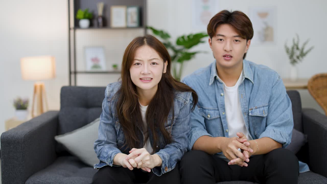 Một người đàn ông và một người phụ nữ châu Á ngồi trên ghế sofa, biểu lộ cảm xúc khi xem TV.