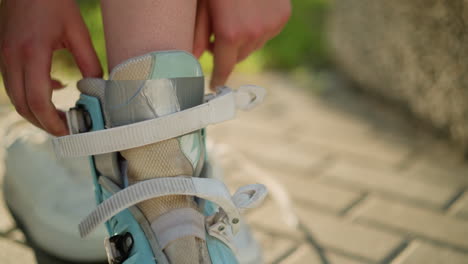 close-up of individual adjusting roller skate straps with both hands, with two white sneakers blurred in background, greenery and sunlight creating soft backdrop, person preparing for skating