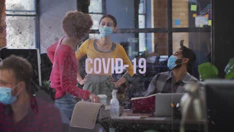 Covid-19-text-against-office-colleagues-wearing-face-mask-laughing-together-at-office
