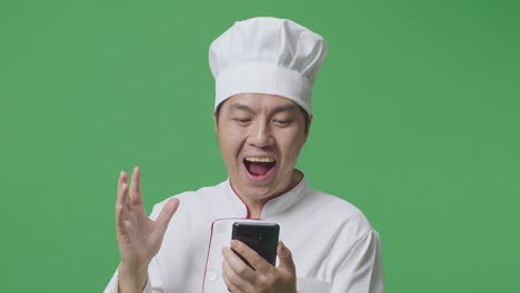 close up de un chef asiático mirando su teléfono inteligente, sonriendo, y diciendo wow mientras está de pie en el estudio de fondo de la pantalla verde