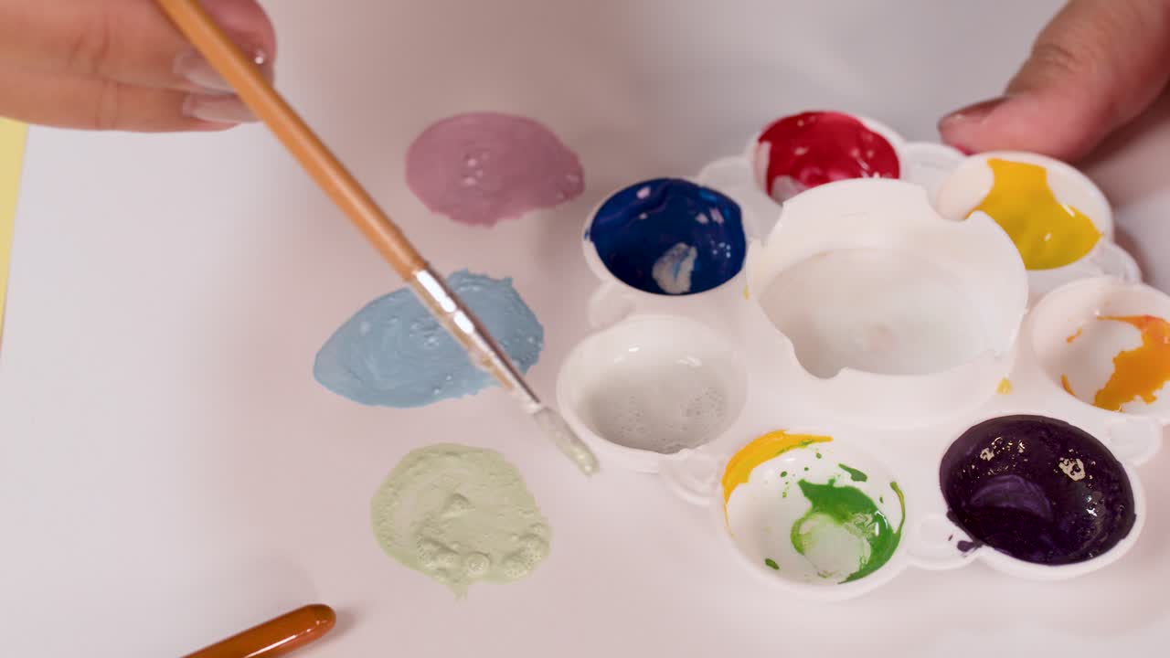 Hand mixes green watercolor paint on palette using brush, bright lighting, overhead camera, creative workspace