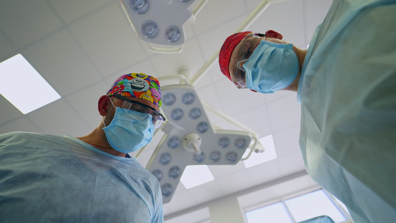 A couple of male doctors in funny hats, masks and glasses talking and consulting during surgery. Low angle view from a patient's spot.