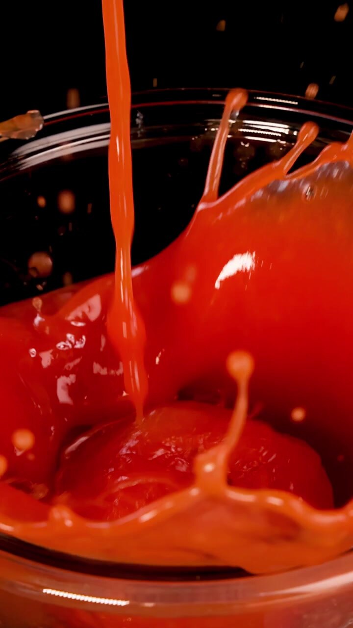 Un pomodoro intero viene fatto cadere in una ciotola di vetro piena di salsa di pomodoro, creando schizzi e bolle sotto una luminosa illuminazione da studio con uno sfondo nero.