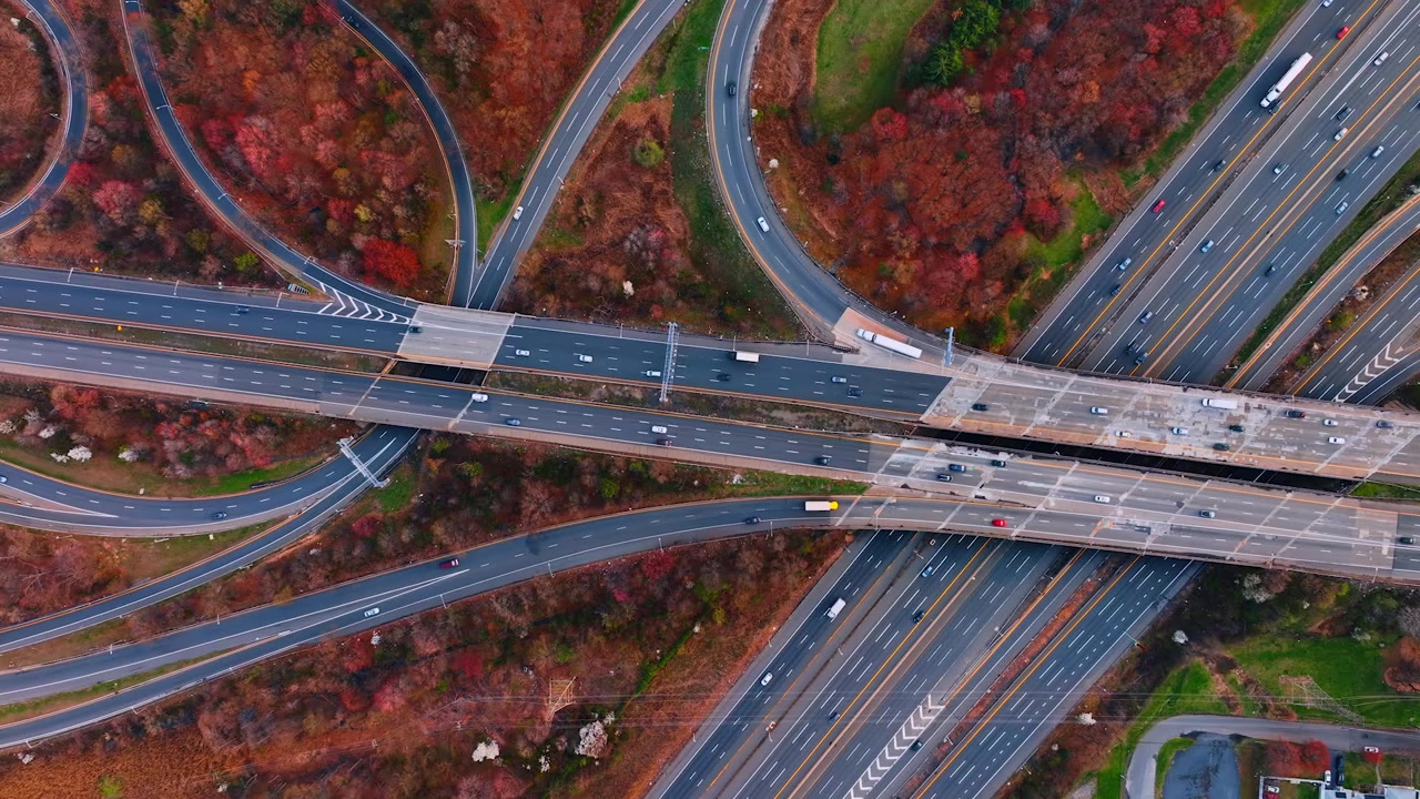 Aerial of a freeway interchange near dense suburban housing, New Jersey. Top-down of multilane lanes and a crossing overpass with autumn trees, homes and a basketball court nearby