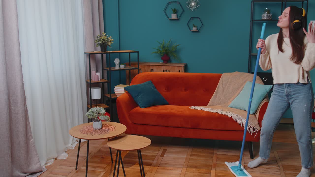 Joyful young woman is mopping floor in apartment living room having fun listening to music at home