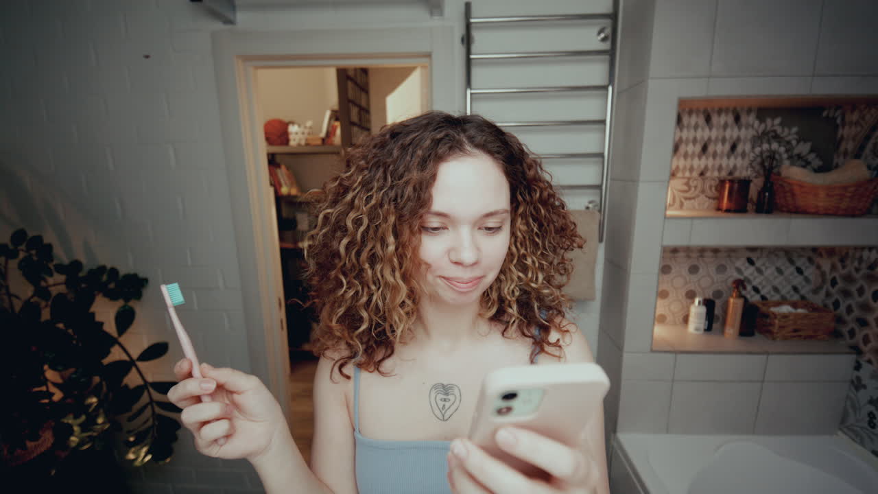 chica de cabello rizado cepillándose los dientes y usando el teléfono inteligente en el baño
