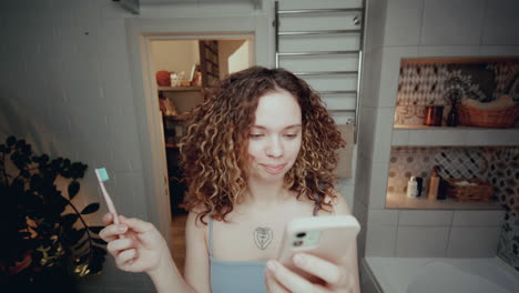 curly-haired girl brushing teeth and using smartphone in bathroom