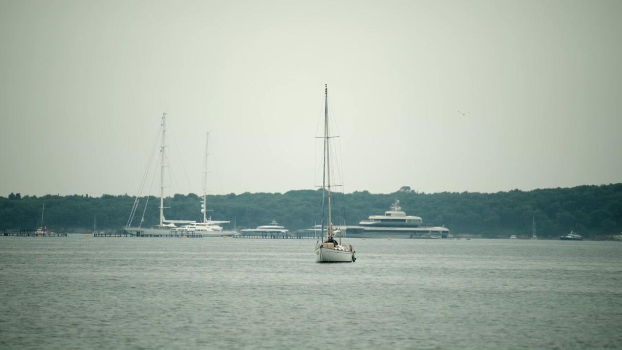 Barca a vela su un mare calmo, che passa accanto a superyacht di lusso ormeggiati lungo una costa boscosa sotto un cielo coperto.